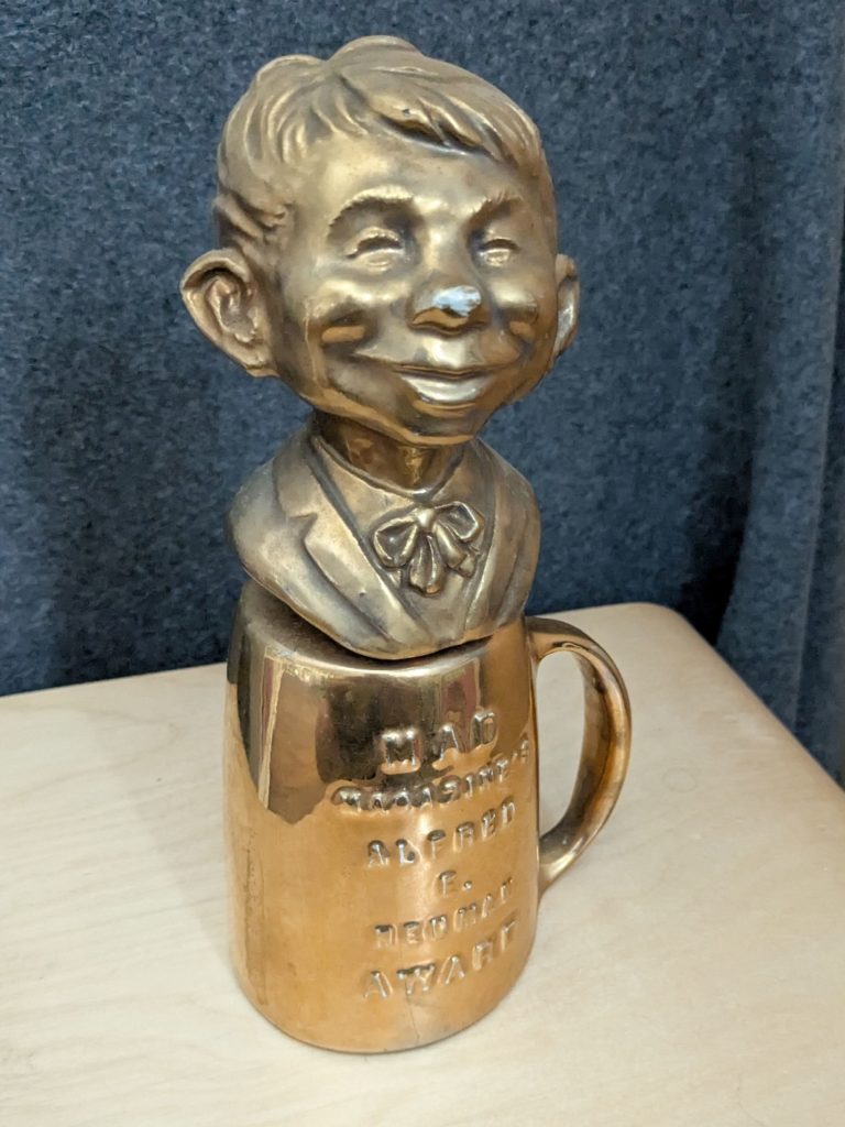 Ein goldfarbener Alfred E. Neuman Award, bestehend aus einer Büste von Alfred E. Neuman, die auf einer umgedrehten Tasse mit der Aufschrift "MAD Magazine's Alfred E. Neuman Award" befestigt ist. Der Award diente als Requisite in TV-Shows und ist ein seltenes Sammlerstück.