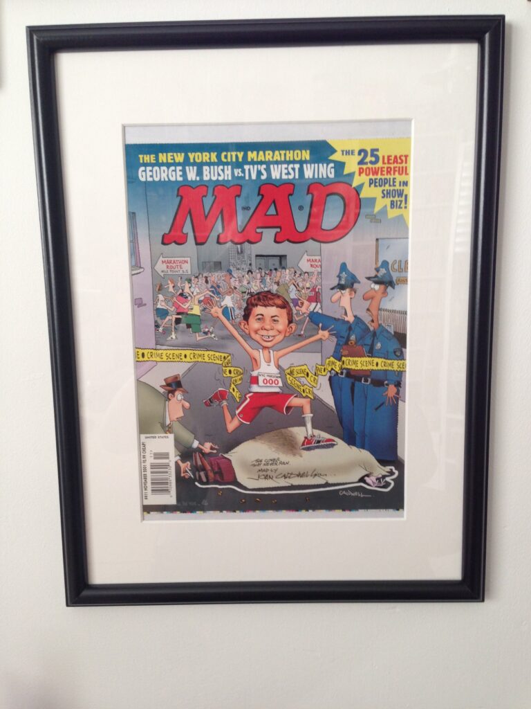 Gerahmtes, unveröffentlichtes Titelbild der US-Ausgabe MAD #411 von John Caldwell. Alfred E. Neuman rennt beim New York Marathon lachend durch ein Polizei-Absperrband an einem Tatort.