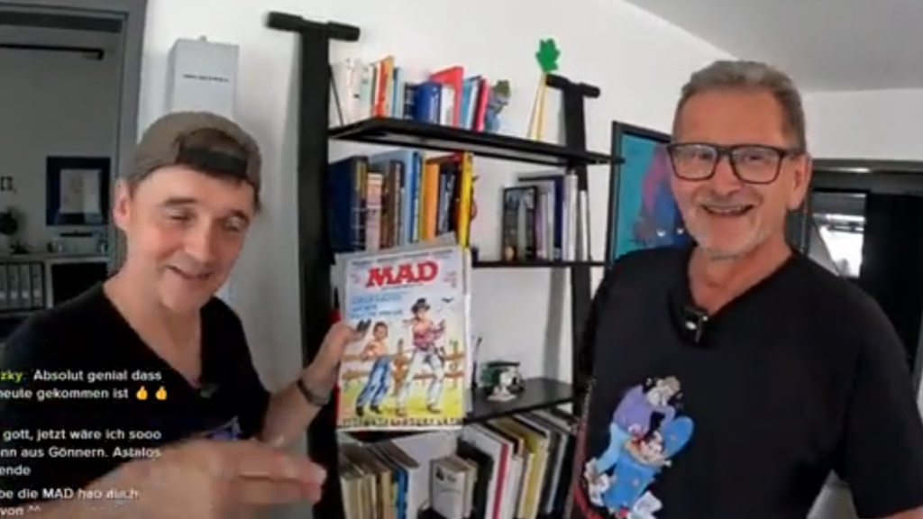 Paddy und der deutsche MAD-Zeichner Ivica Astalos stehen nebeneinander vor einem Regal. Paddy hält lächelnd ein klassisches MAD-Magazin in die Kamera.
