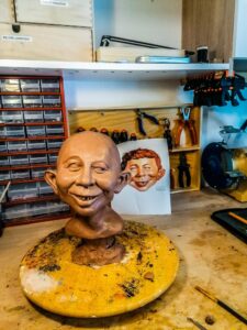 Rohes Tonmodell einer Alfred E. Neuman Büste auf einem gelben Drehteller in einer Werkstatt, dahinter ein Ausdruck der Vorlage.