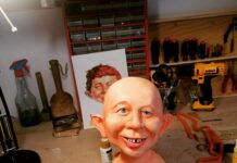 Abgeformter, noch haarloser Kopf der Alfred E. Neuman Büste während der realistischen Bemalung mit blauen Augen, Sommersprossen und Zahnlücke, davor eine Farbpalette.