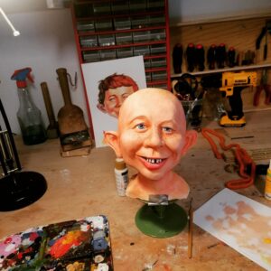 Abgeformter, noch haarloser Kopf der Alfred E. Neuman Büste während der realistischen Bemalung mit blauen Augen, Sommersprossen und Zahnlücke, davor eine Farbpalette.