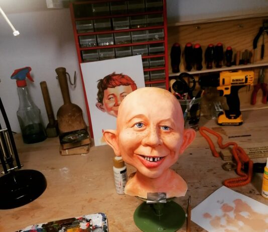Abgeformter, noch haarloser Kopf der Alfred E. Neuman Büste während der realistischen Bemalung mit blauen Augen, Sommersprossen und Zahnlücke, davor eine Farbpalette.
