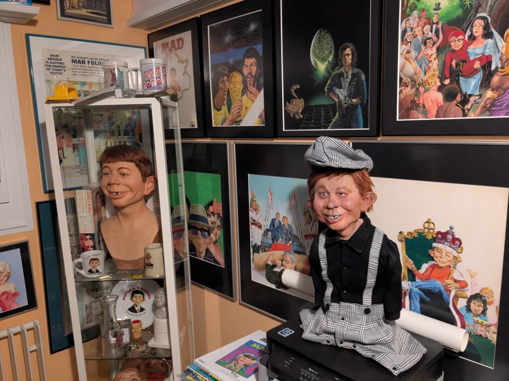 Die handgefertigte Alfred E. Neuman Büste von Pierre Schrader im Hahnentritt-Outfit steht in einem Sammlerzimmer neben einer Glasvitrine mit der seltenen Warner Bros. Büste von Gary Mirabelle, umgeben von gerahmten MAD-Originalkunstwerken.
