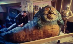 Bildhauer Pierre Schrader lehnt in seiner Werkstatt neben einer lebensgroßen Skulptur von Jabba the Hutt, im Hintergrund stehen C-3PO und Han Solo in Karbonit.